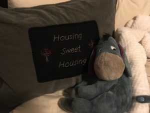 A gray pillow with a black section embroidered with "Housing sweet housing" sits behind a stuffed animal Eeyore
