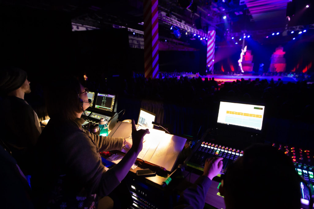 A stage manager has one finger up in standby as acrobat lower to the stage