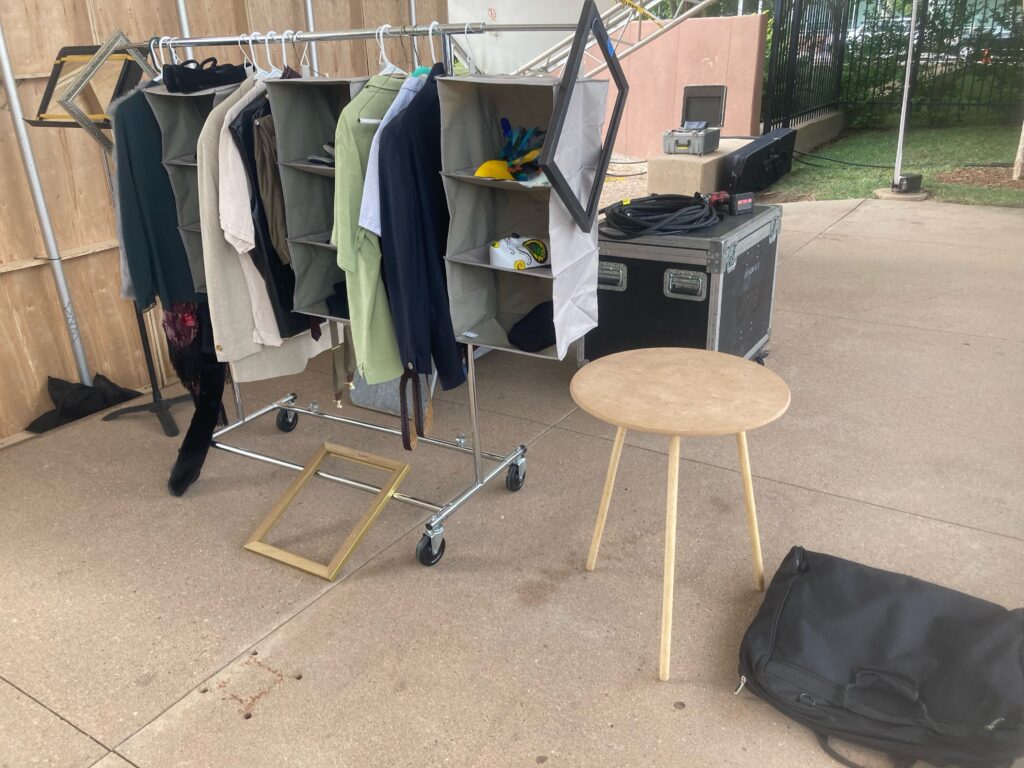 A costume rack holds costumes and hanging shelves for props. To the right is a collapsible round table and on the floor next to it a garment bag. To the left is a music stand with a mirror.