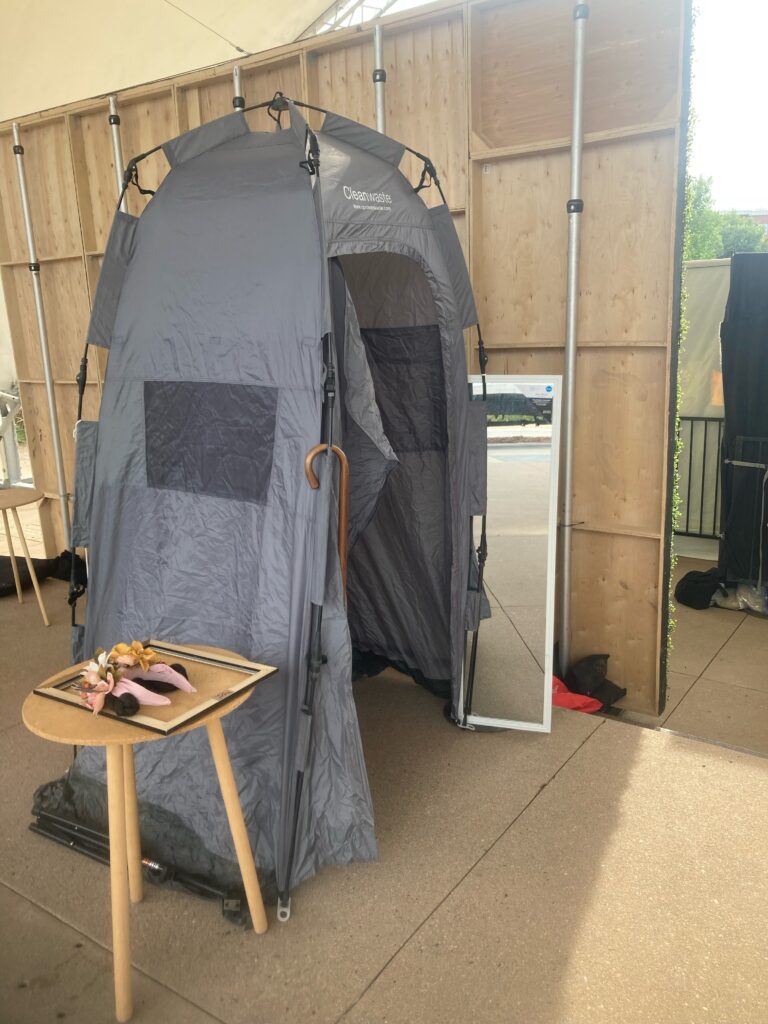 A tall skinny gray tent stands backstage next to a tall mirror, along side a round table with props on it.