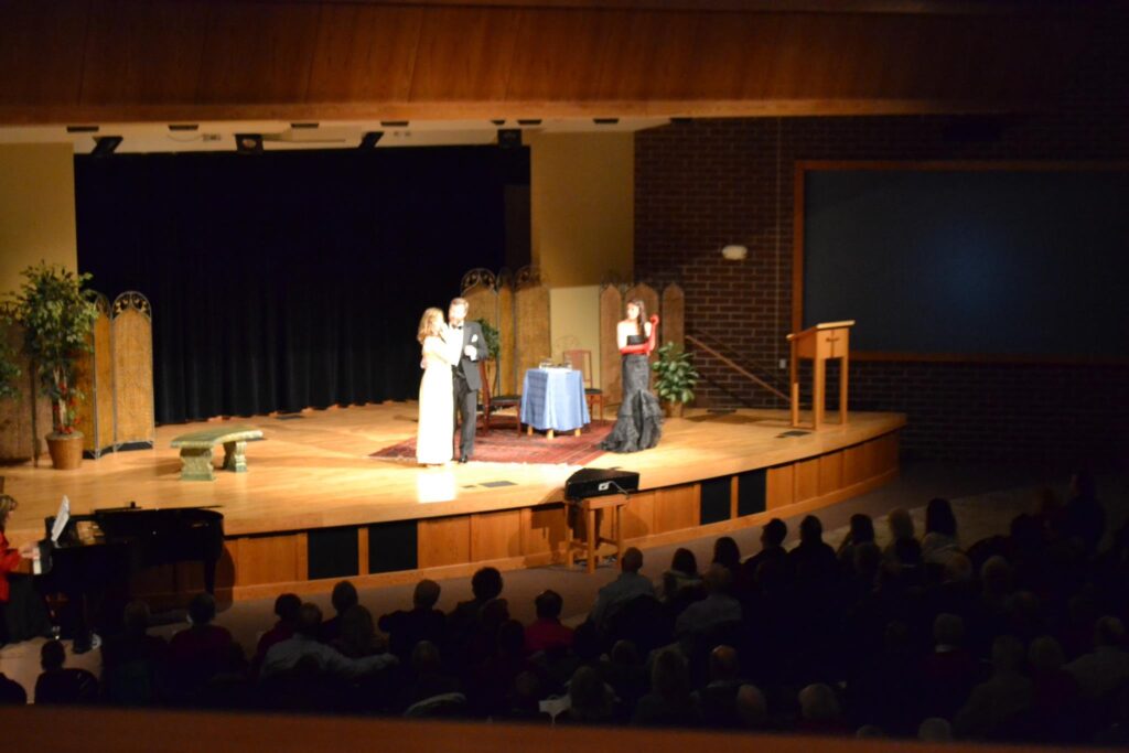 a wide stage has been transformed with 4 wicker screens, several fake plants and trees, and a tapestry rug.