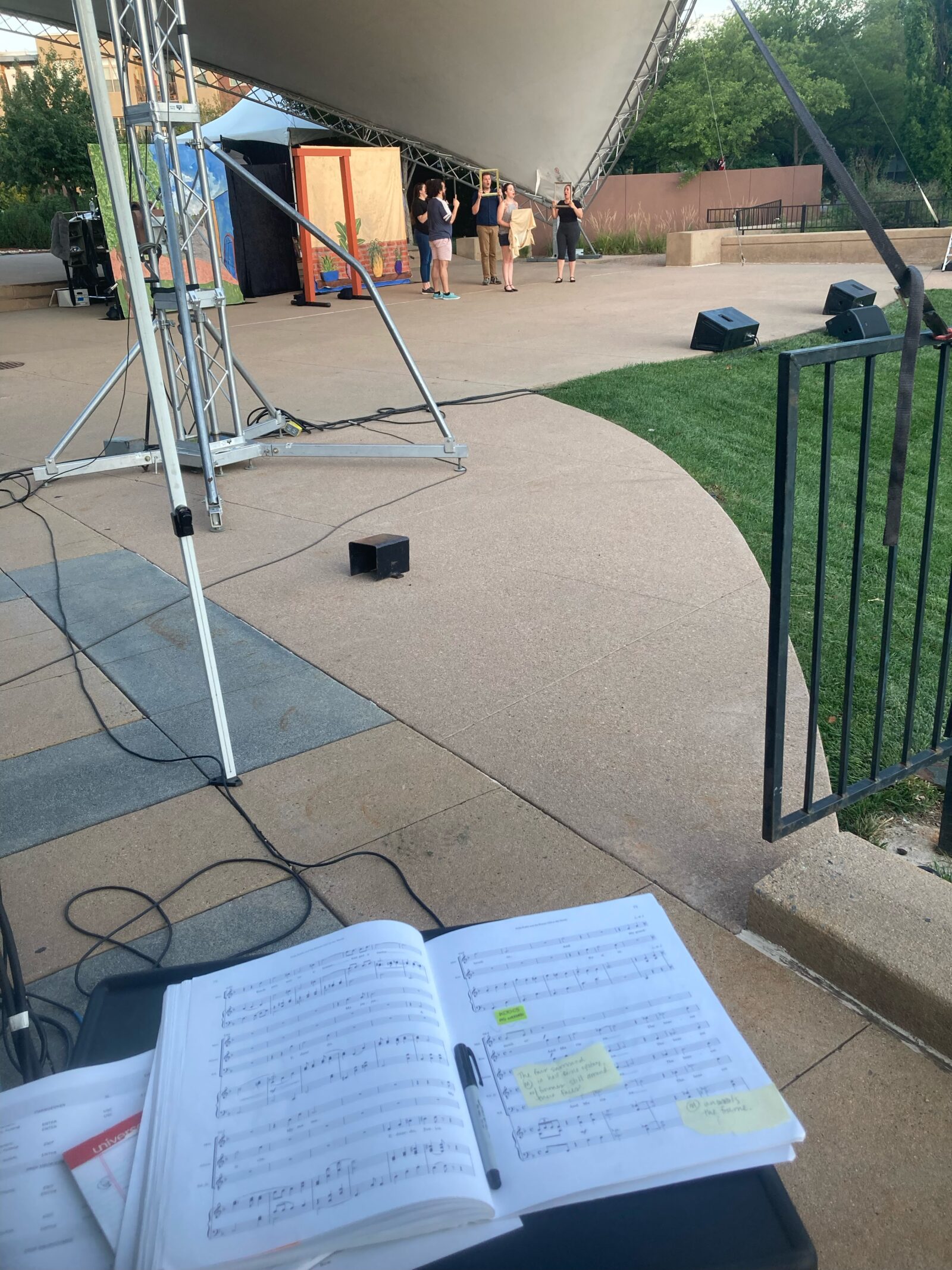 The view from the stage manager’s position during an outdoor opera tech rehearsal. The score is sitting in front, covered in post-it notes. On stage, five actors hold up picture frames in front of colorful drops and lighting equipment.