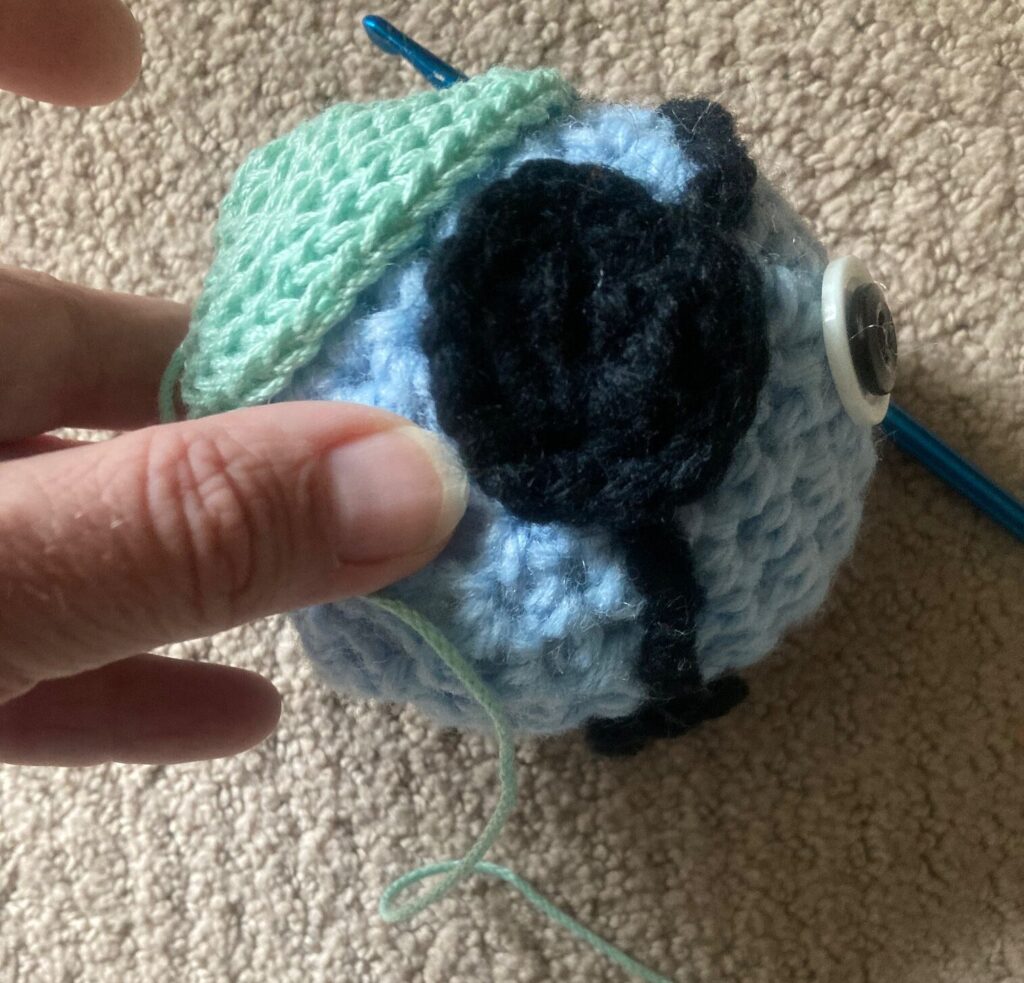 A blue crocheted ball with a black headset AKA a SMeep, is shown with another smaller crocheted green piece on the back, which looks a bit like a yarmulke