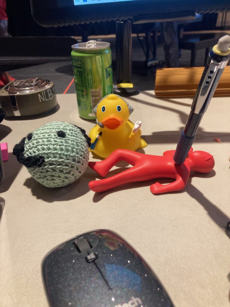 A stage manager's desk is shown, with three mascots: a crocheted SMeep (green ball with black yarn headset), a Stage Manager Rubber Duck, and a pencil holder that looks like the pencil has stabbed a red humanlike figure