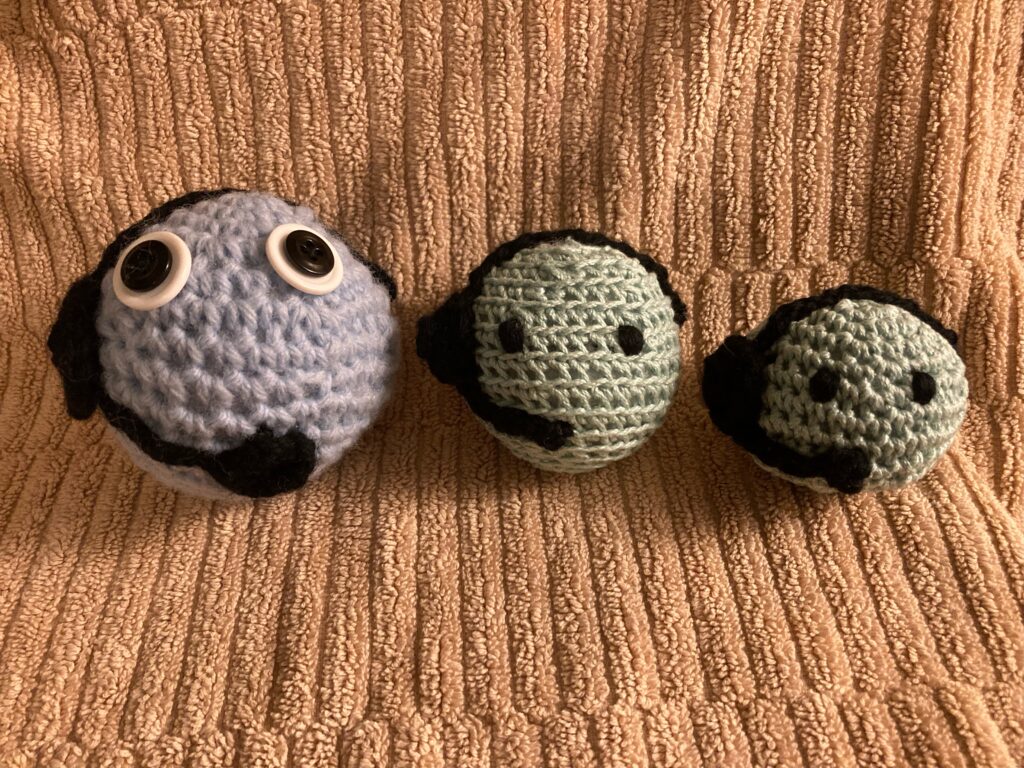 Three crocheted balls sit on a brown textured towel. Each of them has eyes and a black headset added. The left one is biggest and blue. The middle is round and green. The third is also green but is flatter in shape than the first two.