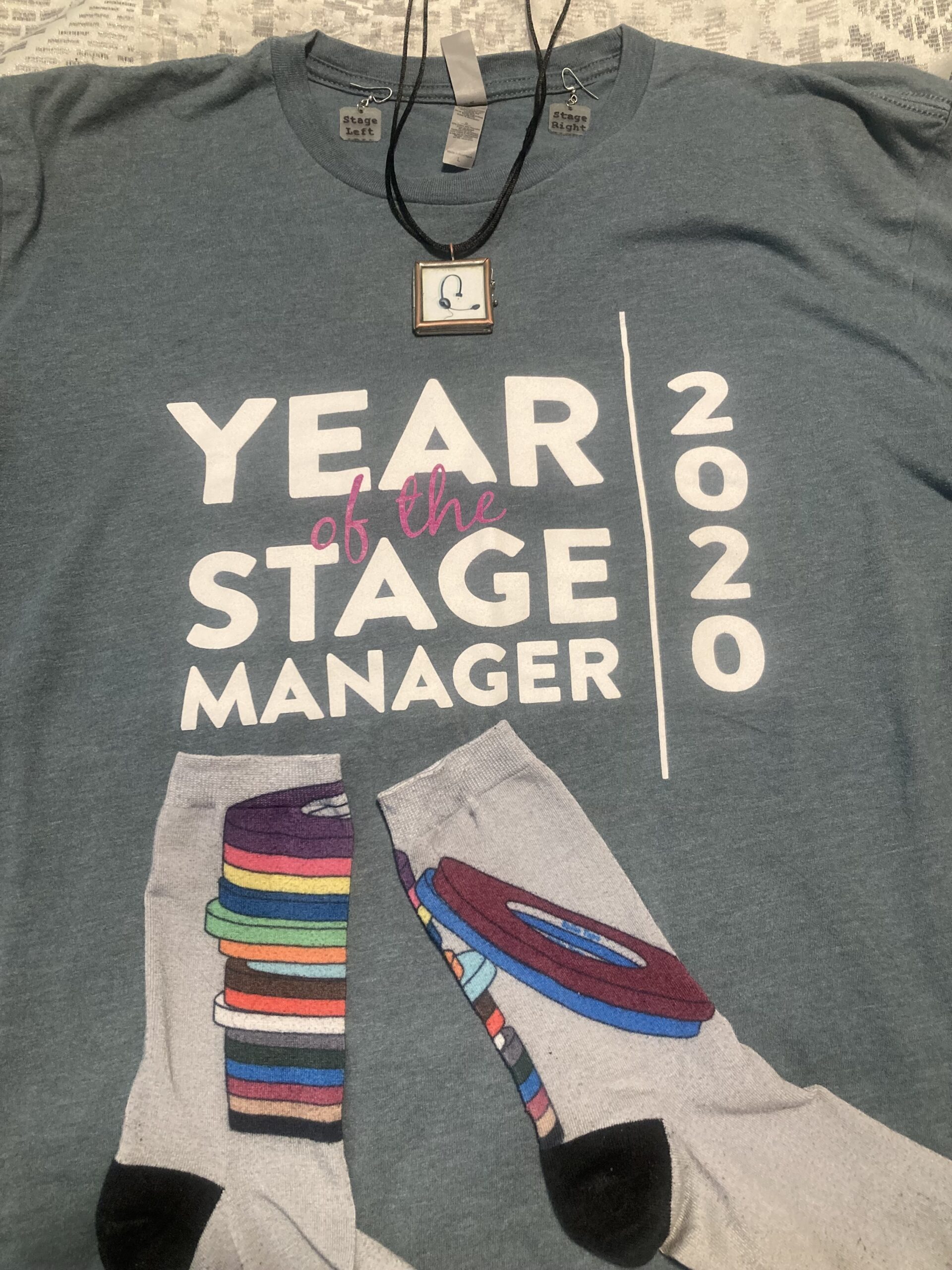 A t-shirt reads Year of the Stage Manager 2023, on top of it are two socks with images of spike tape rolls. Placed on top of the shirt are also a stage management headset pendant necklace and earrings that say Stage Left and Stage Right