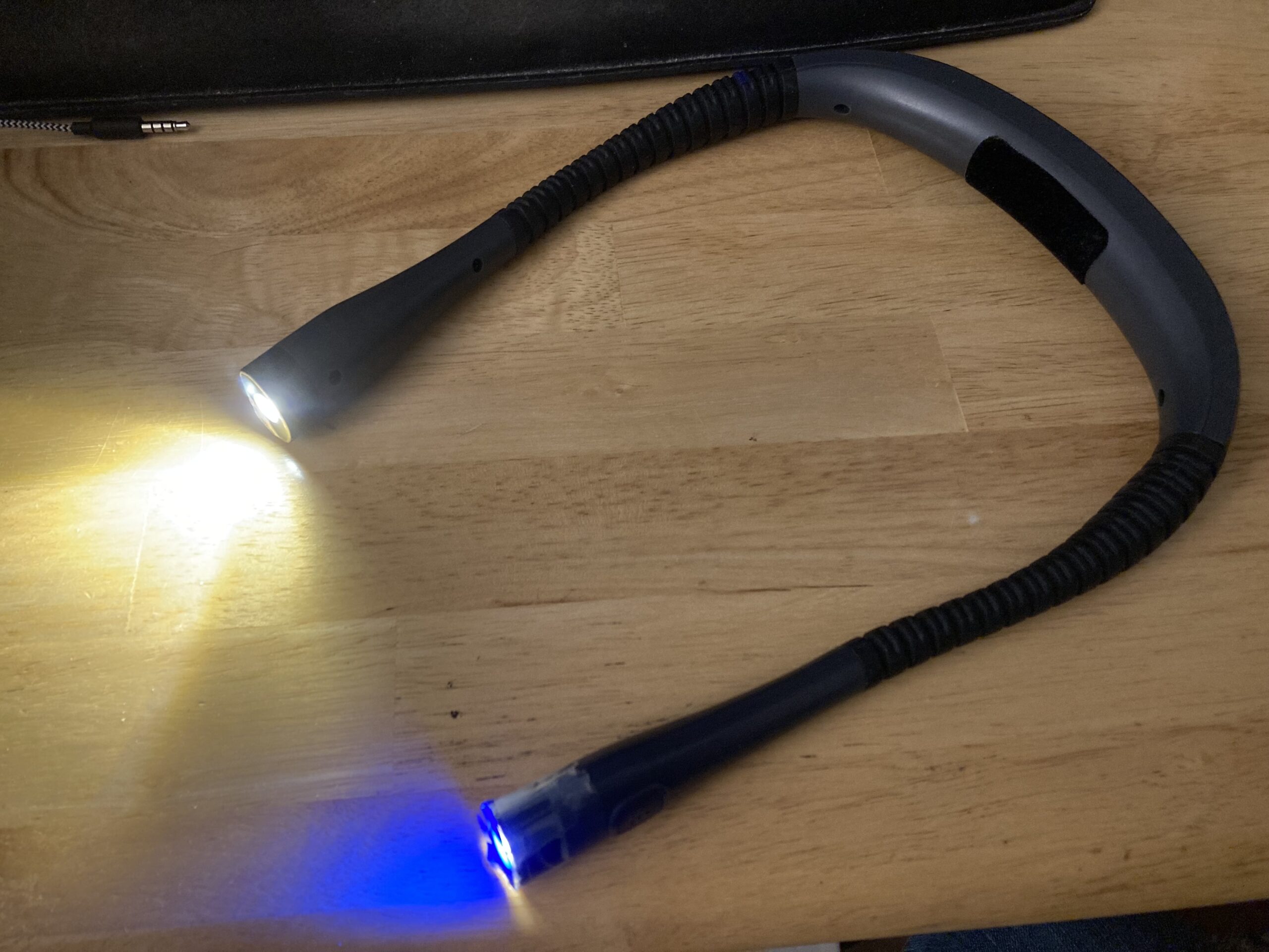 A closeup of a necklight flashlight sitting on a table. Both ends have lights turned on, one with a blue tint.