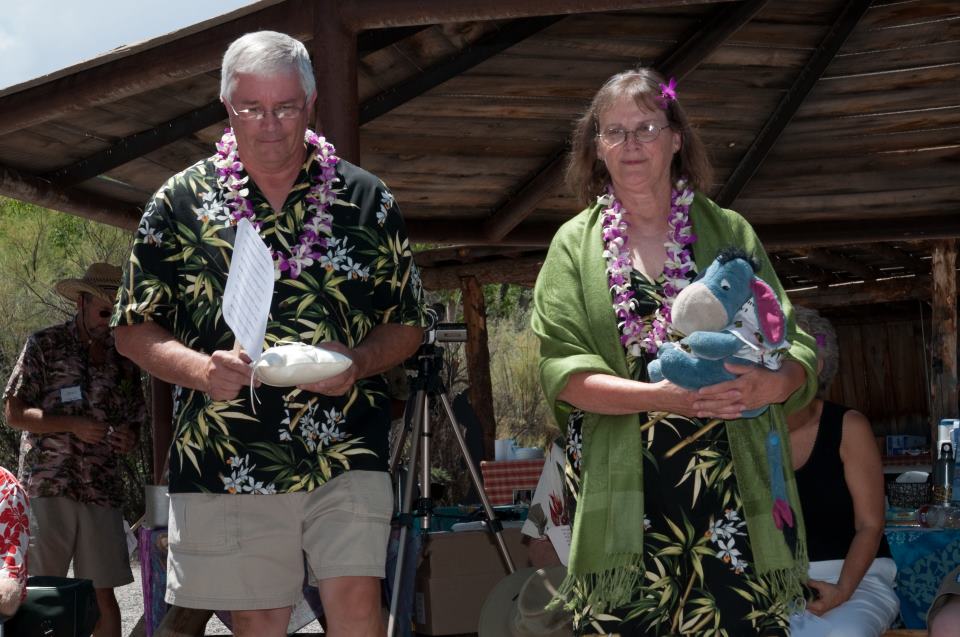 A man and a woman in Hawaiian style clothing and leis head towards the camera. The woman is holding a stuffed blue Eeyore, than man a pillow.