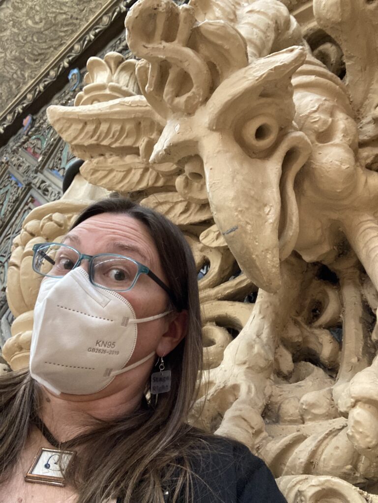 A Woman with a face mask looks with a quizzical face next to a bird-type statue on the wall. She is wearing a headset necklace.