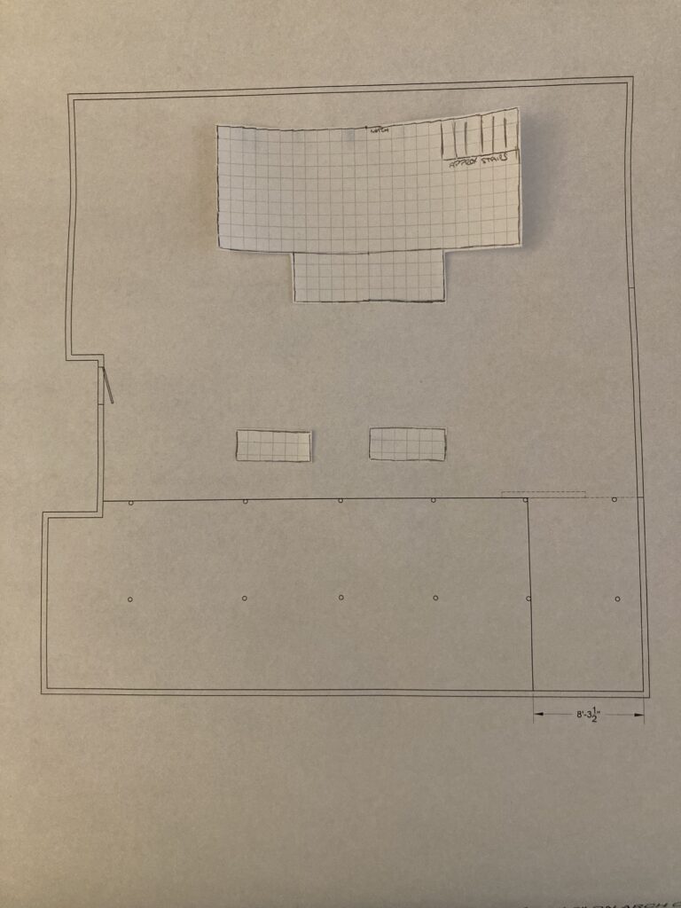 A rehearsal hall groundplan is shown with cut out pieces of graph paper on top of it to indicate a platform and tables.