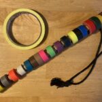 A spike stick is pictured, with 19 colors of spike tape rolled around a dowel. The dowel has a hole drilled towards one end, with black tieline laced through and tied in a knot as a loop. Above and to the left is a single roll of yellow spike tape.