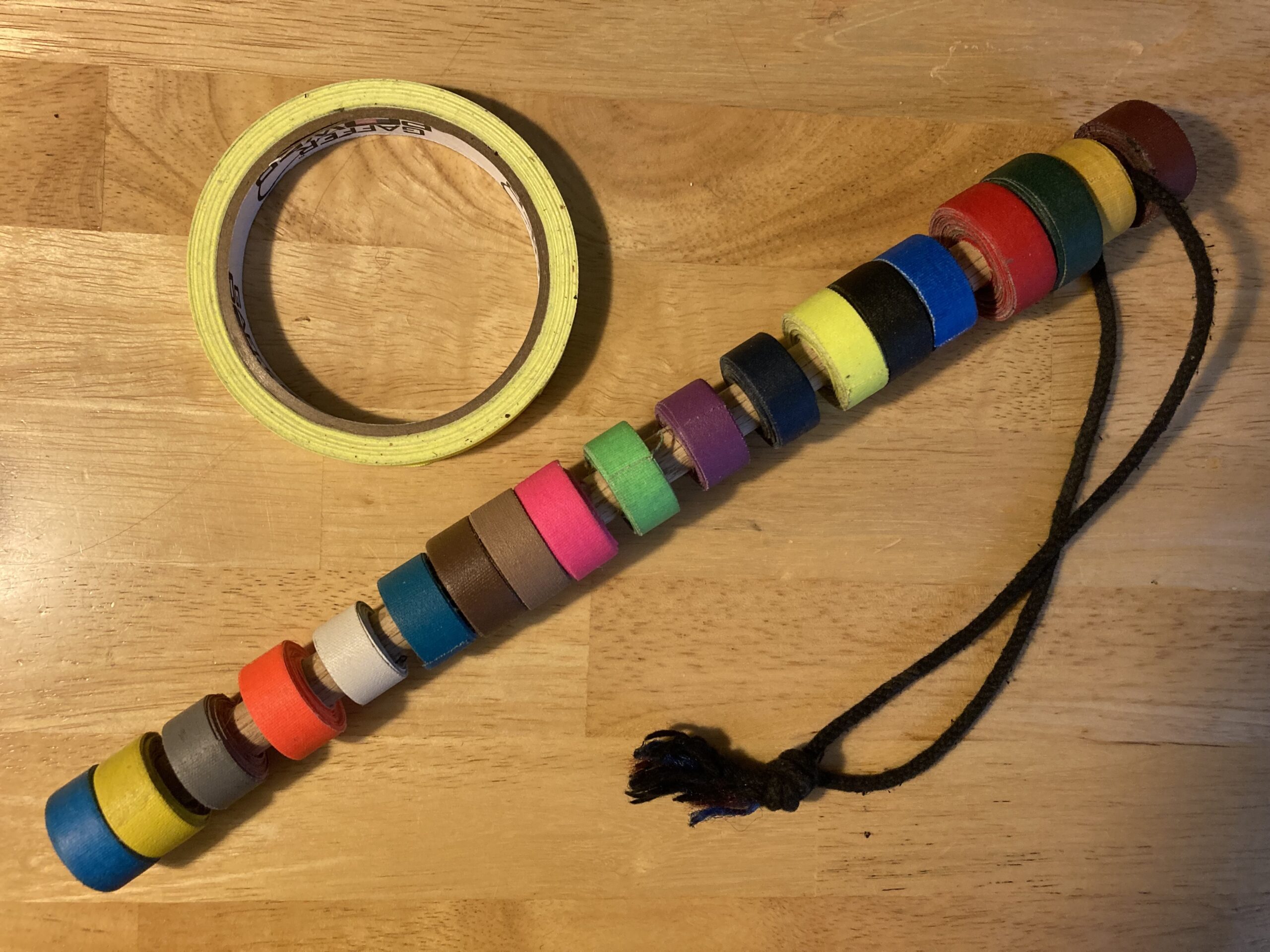 A spike stick is pictured, with 19 colors of spike tape rolled around a dowel. The dowel has a hole drilled towards one end, with black tieline laced through and tied in a knot as a loop. Above and to the left is a single roll of yellow spike tape.