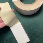 White gaff tape is layered around a plastic hotel key for storage. The end of the tape is loose in someone's hand, and a roll of the same gaff sits in the background, all on a green velvet background.