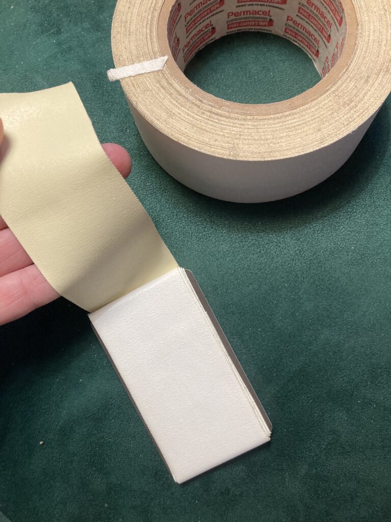 White gaff tape is layered around a plastic hotel key for storage. The end of the tape is loose in someone's hand, and a roll of the same gaff sits in the background, all on a green velvet background.